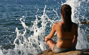 Kvinne sitter ved havet i lotusstilling og sjøen spruter foran henne