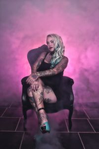 Kvinne med kroppen full av tatoveringer sittende i en stol omringet av røyk