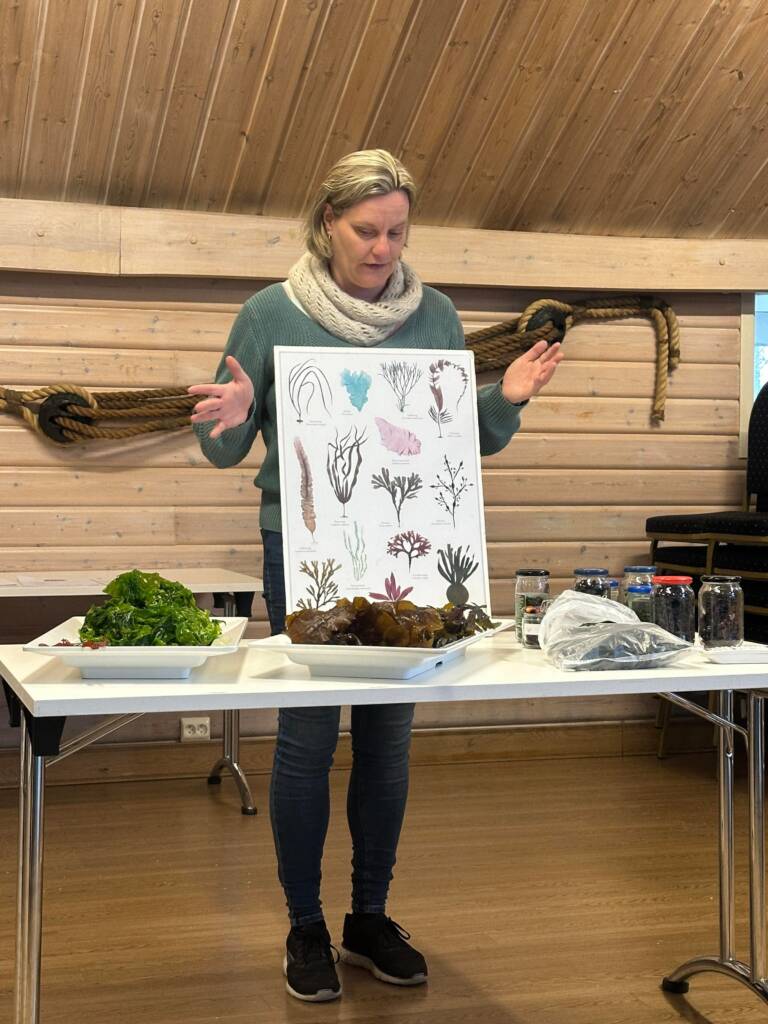 Melanie Endemann holder opp en plakat med tang- og taresorter, ved et bord fylt med ferske produkter.