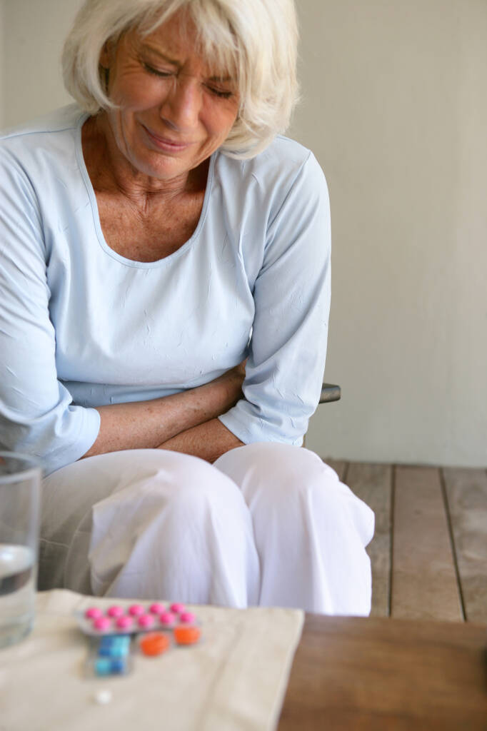 Eldre kvinner som lager grimaser og holder seg på magen i smerter. Noen fargerike piller ligger på bordet ved siden av.