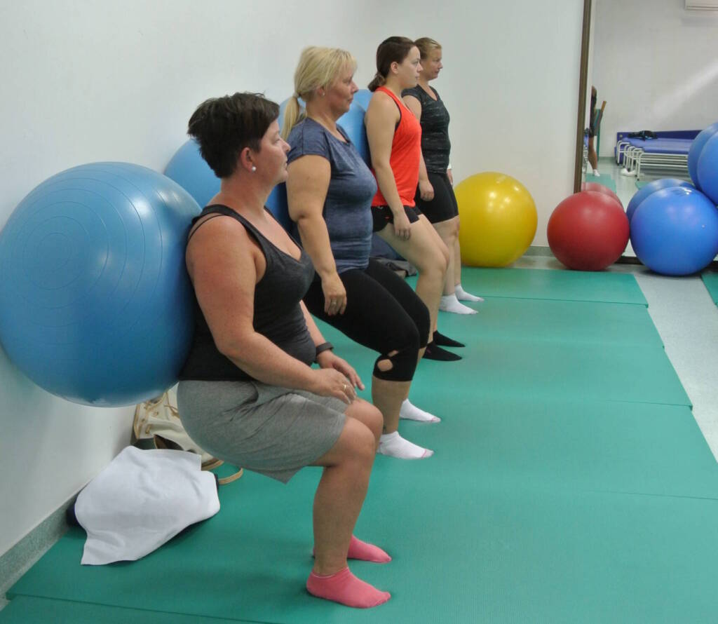 Fire personer står i treningssal og lener seg på en stor pilatesball mot veggen, som de fører opp og ned ved å bøye knærne